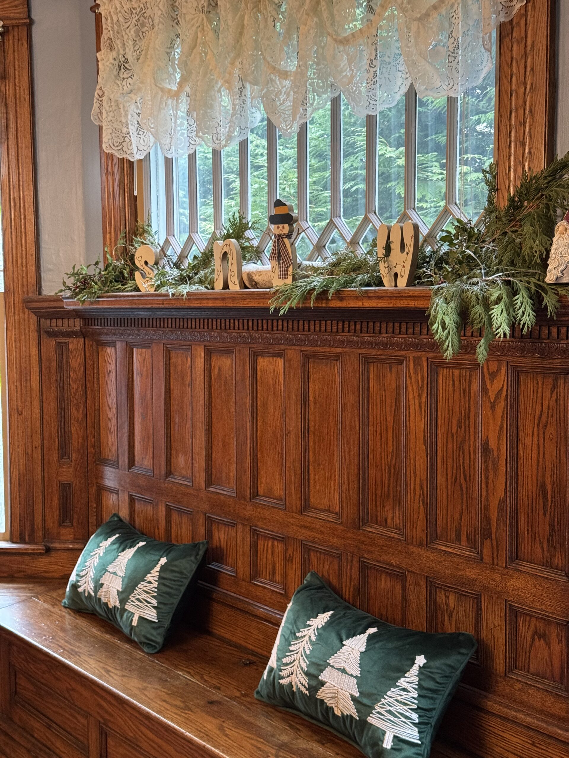 Cozy winter scene with embroidered green pillows on a wooden bench, decorated with snow-themed accents, including wooden letters and greenery.