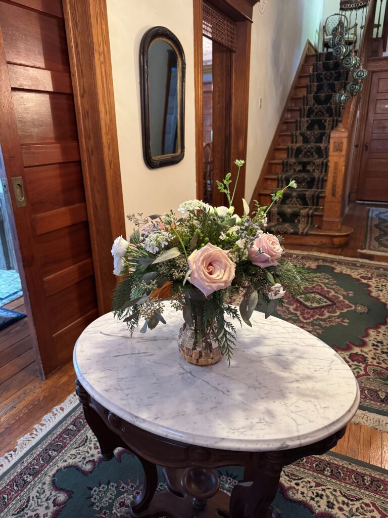 A vase of pastel roses and greenery sits on a marble-topped table in a warmly lit hallway, featuring wooden trim and a staircase in the background.
