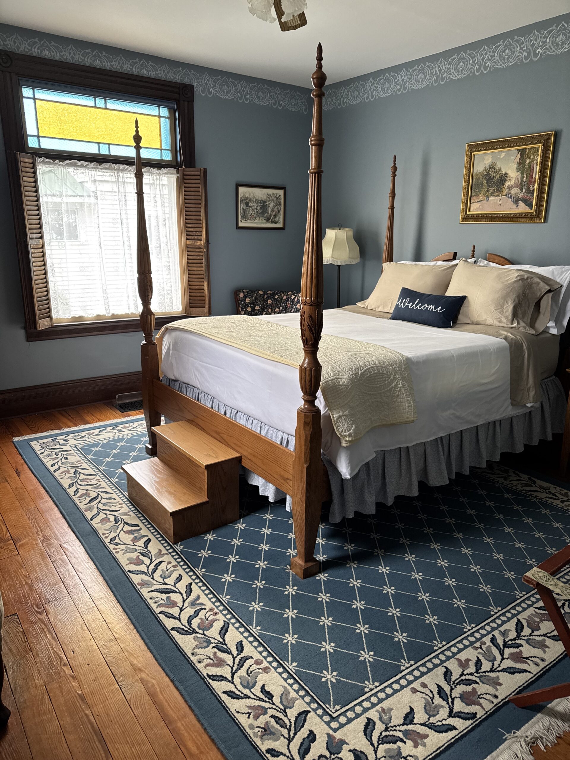 Elegant bedroom featuring a four-poster bed with steps, a patterned rug, and a vintage color palette. The stained glass window adds charm.