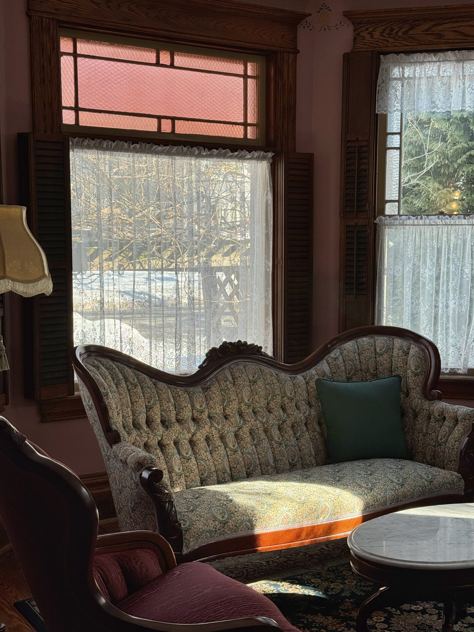 Victorian-style living room featuring a tufted green-patterned sofa with a green cushion, illuminated by natural sunlight through lace curtains.