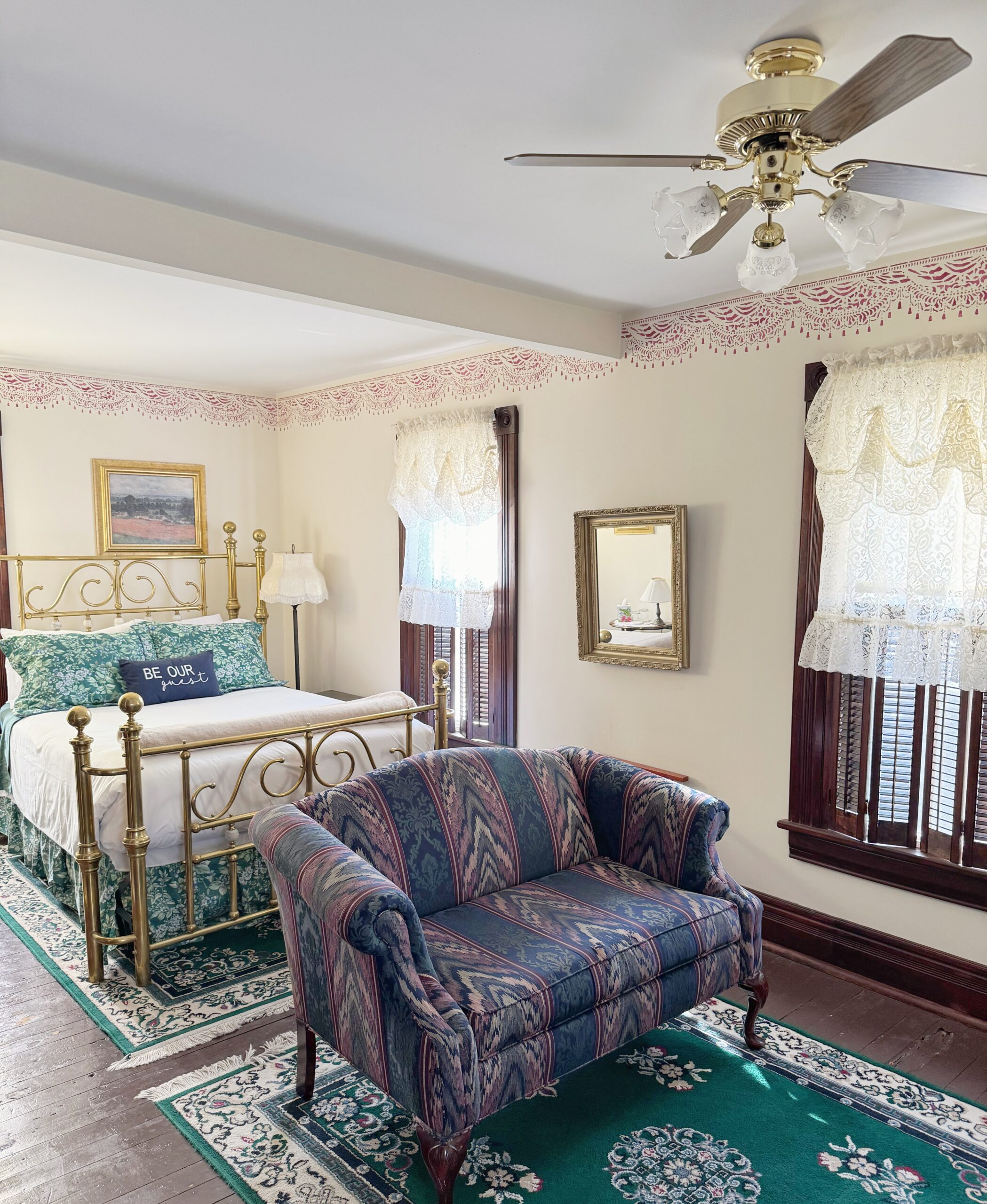 Cozy bedroom featuring a brass bed with floral bedding, a vintage-style loveseat, and intricate lace curtains. Ideal for comfort and relaxation.