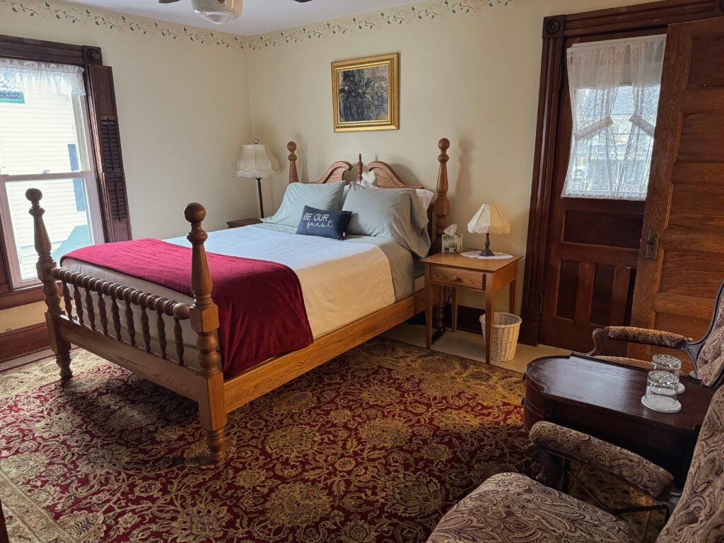 Victorian guest room with a four-poster bed, pale blue bedding, red throw, oriental rug, lace-curtained windows, dark wood trim, vintage lamps, armchairs, and a wicker wastebasket.