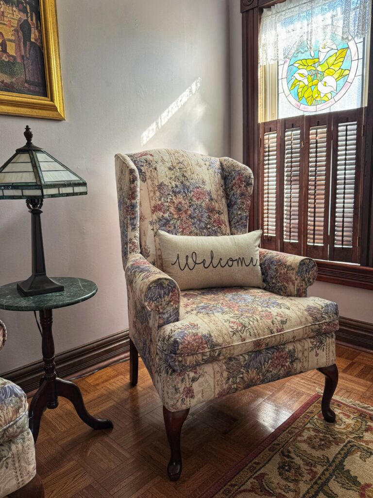 A floral-patterned armchair with a "Welcome" pillow beside a stained glass window and a small lamp on a side table, creating a cozy atmosphere.