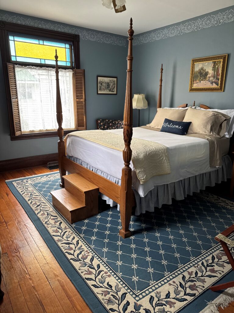 Elegant bedroom featuring a four-poster bed with steps, a patterned rug, and a vintage color palette. The stained glass window adds charm.