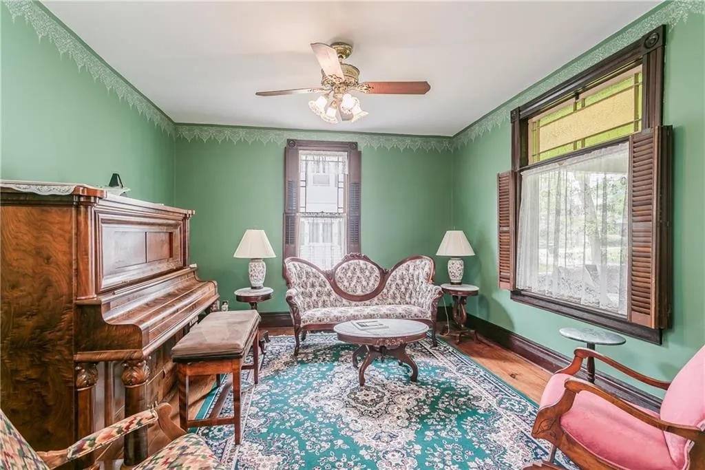 Victorian-style living room featuring a grand piano, ornate seating, and rich green walls, illuminated by natural light from large windows.